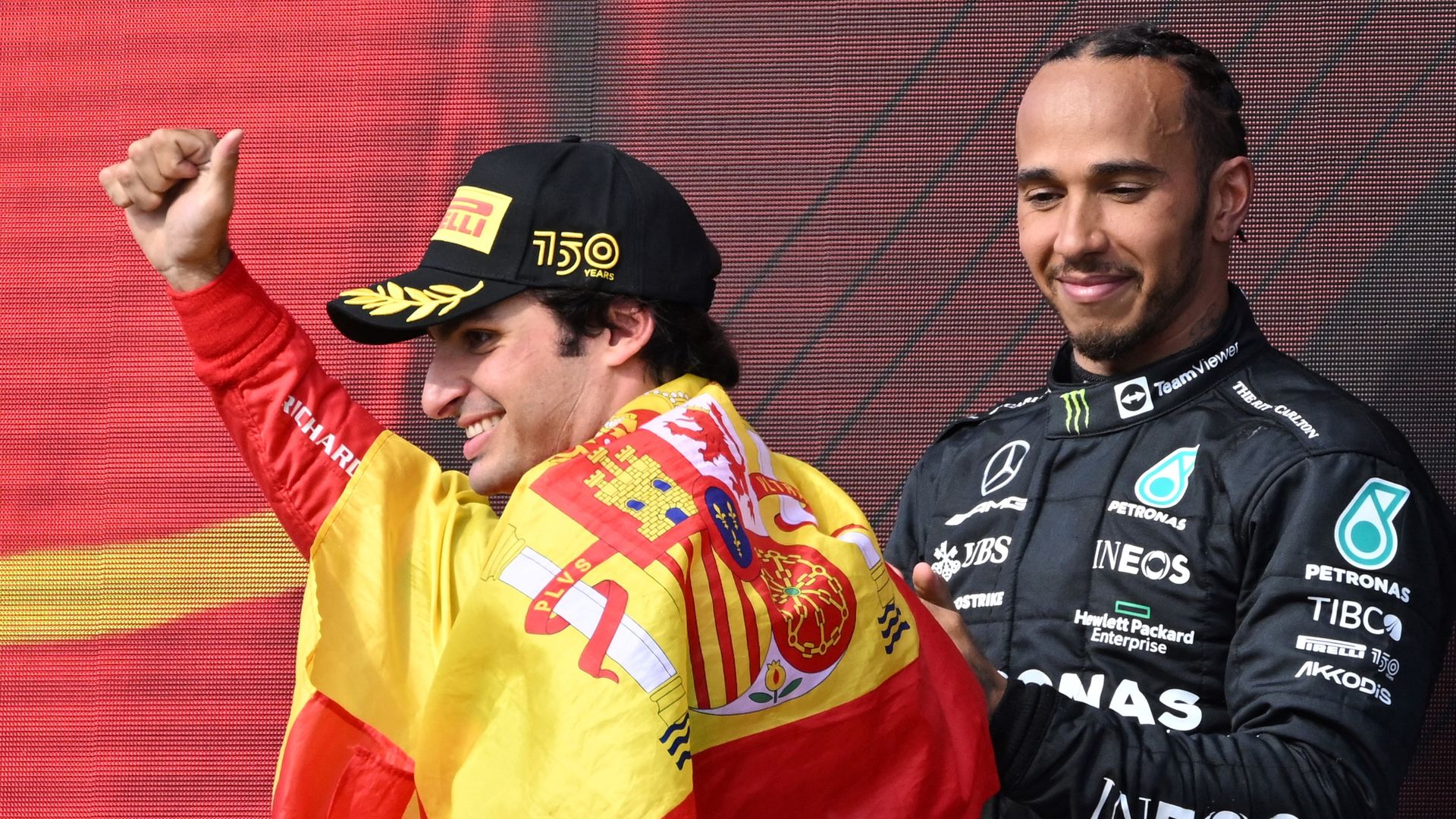 Carlos Sainz, draped in a Spanish flag, celebrates as Lewis Hamilton looks on