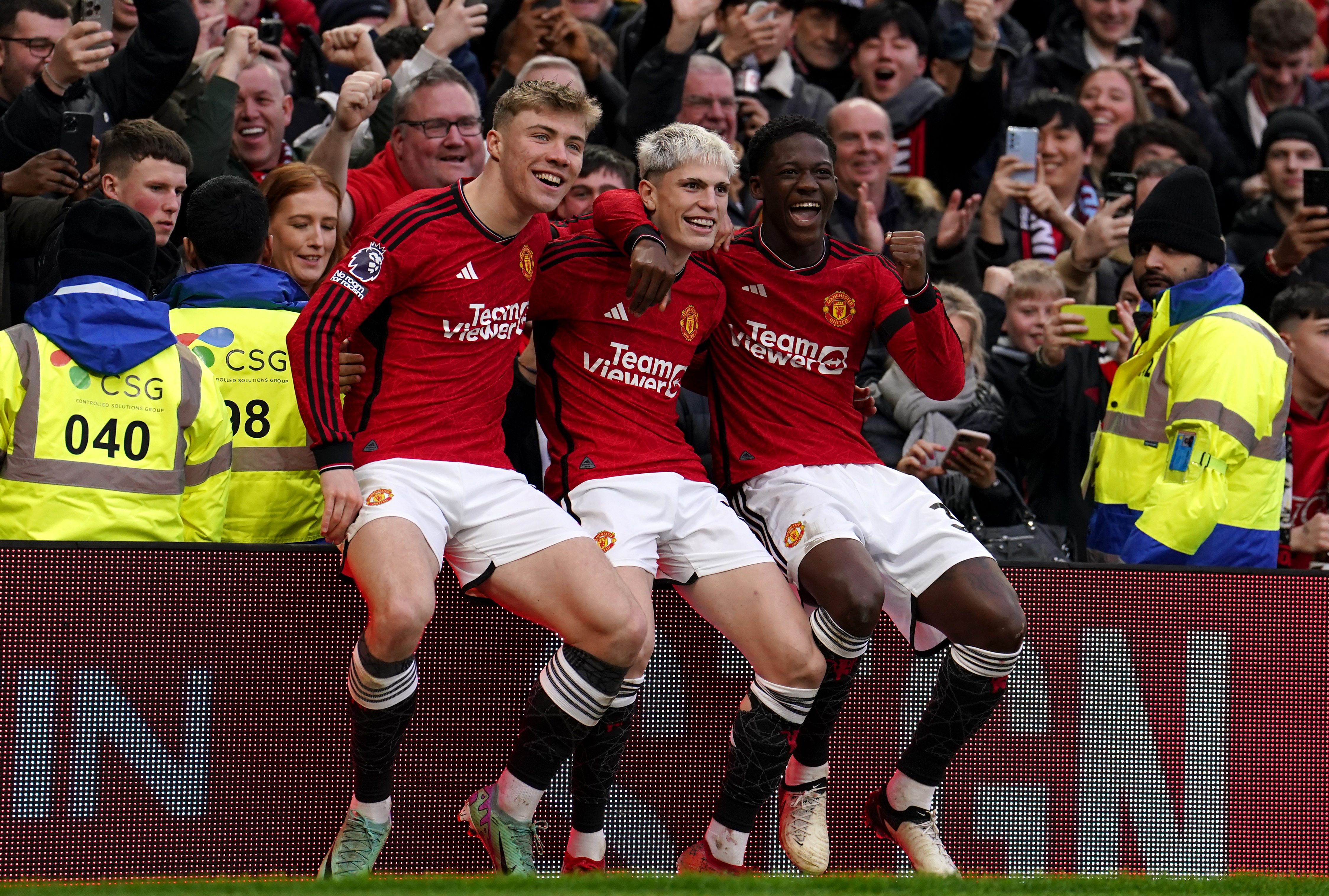Alejandro Garnacho celebrates after scoring against West Ham, sitting on an advertising hoarding flanked by Rasmus Hojlund, left, and Kobbie Mainoo, right