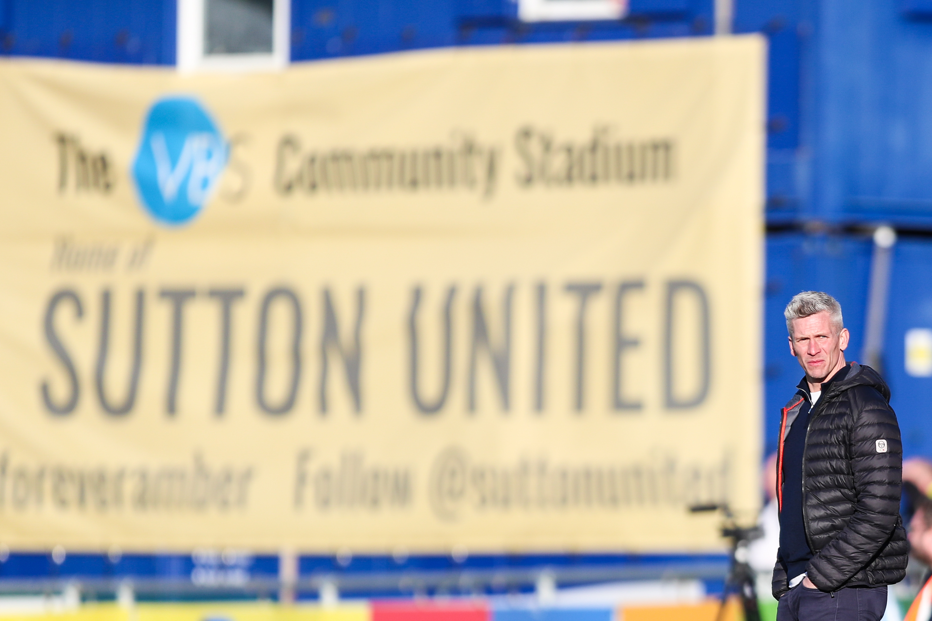 Steve Morison on the touchline in front of a large banner reading 'The VBS Community Stadium, home of Sutton United'